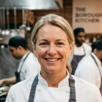 An image of Charlotte Hayes, Kitchen Manager from London. She is smiling and in her kitchen restaurant. In the background are her staff working.